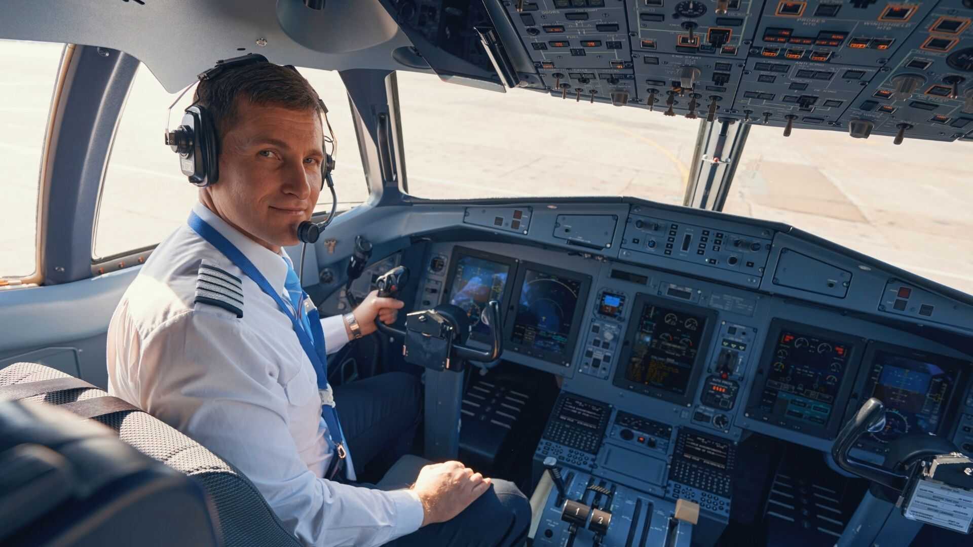 Airline captain in a white uniform and headset seated at the controls of a sunlit modern aircraft cockpit.